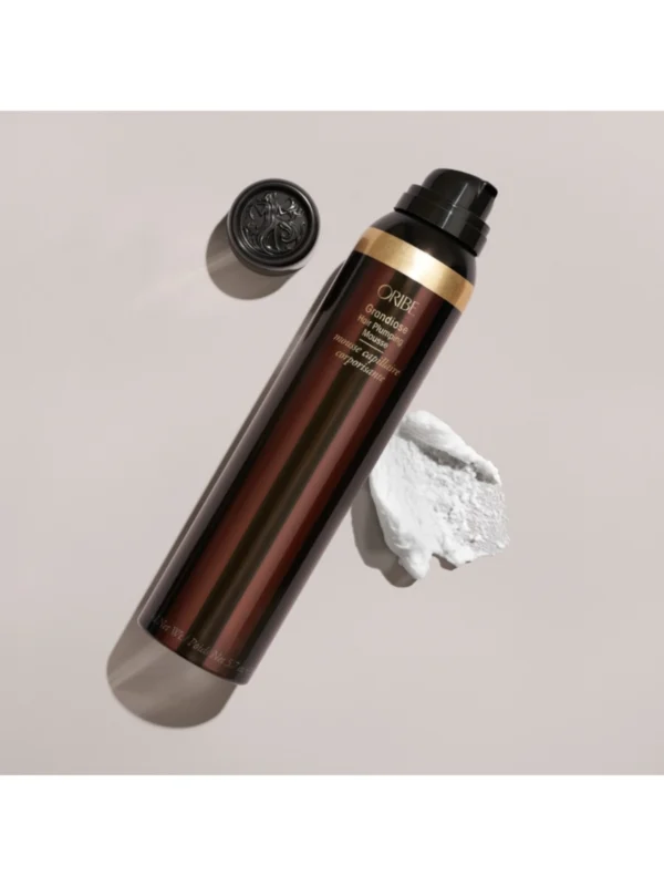 Vista del envase Oribe Grandiose Hair Plumping Mousse junto a una muestra de su textura para peinado profesional