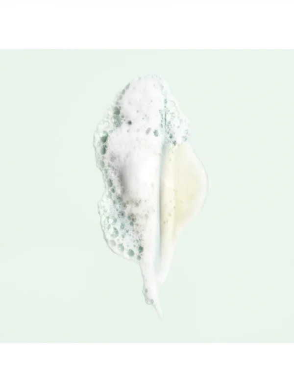 Detalle de la textura en gel y la espuma blanca de Oribe Shampoo For Moisture And Control sobre fondo verde claro