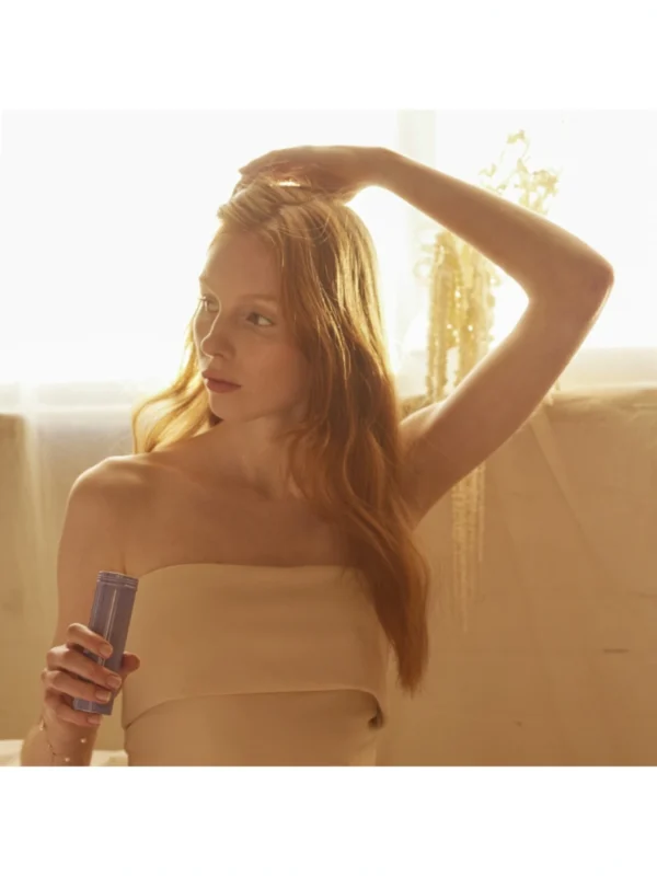 Modelo femenina sosteniendo el envase lila de Serene Scalp Dry Shampoo Powder mientras se arregla el cabello frente a una ventana con luz cálida.