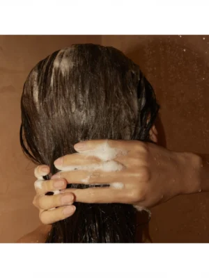 Manos masajeando con espuma la nuca de una persona con cabello castaño mojado durante el lavado