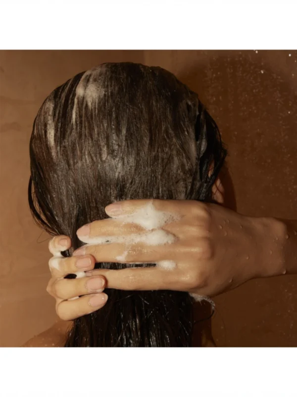 Manos masajeando con espuma la nuca de una persona con cabello castaño mojado durante el lavado