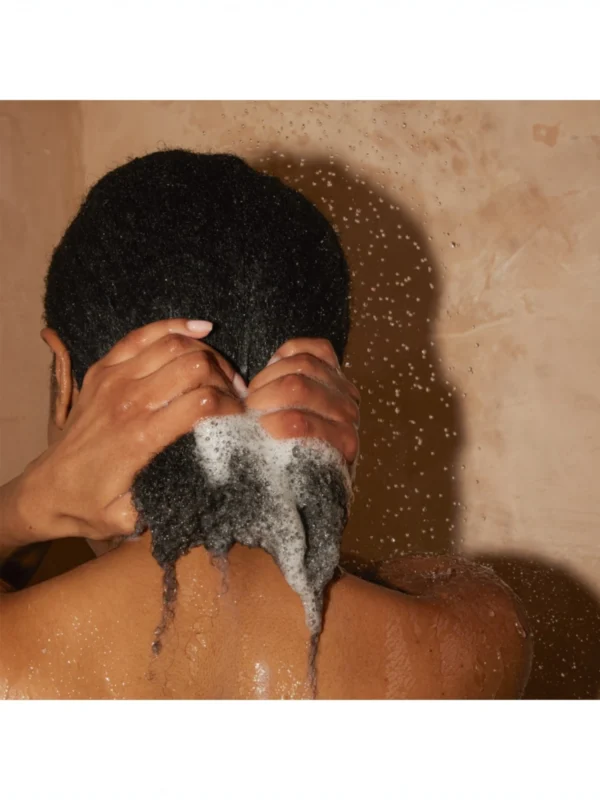 Persona de espalda con cabello oscuro muy rizado aplicando el champú bajo el agua de la ducha