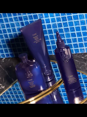 Conjunto de tres productos de la línea Supershine de Oribe (Champú, Acondicionador y Mirror Rinse) presentados sobre una bandeja dorada con fondo de azulejos azules.